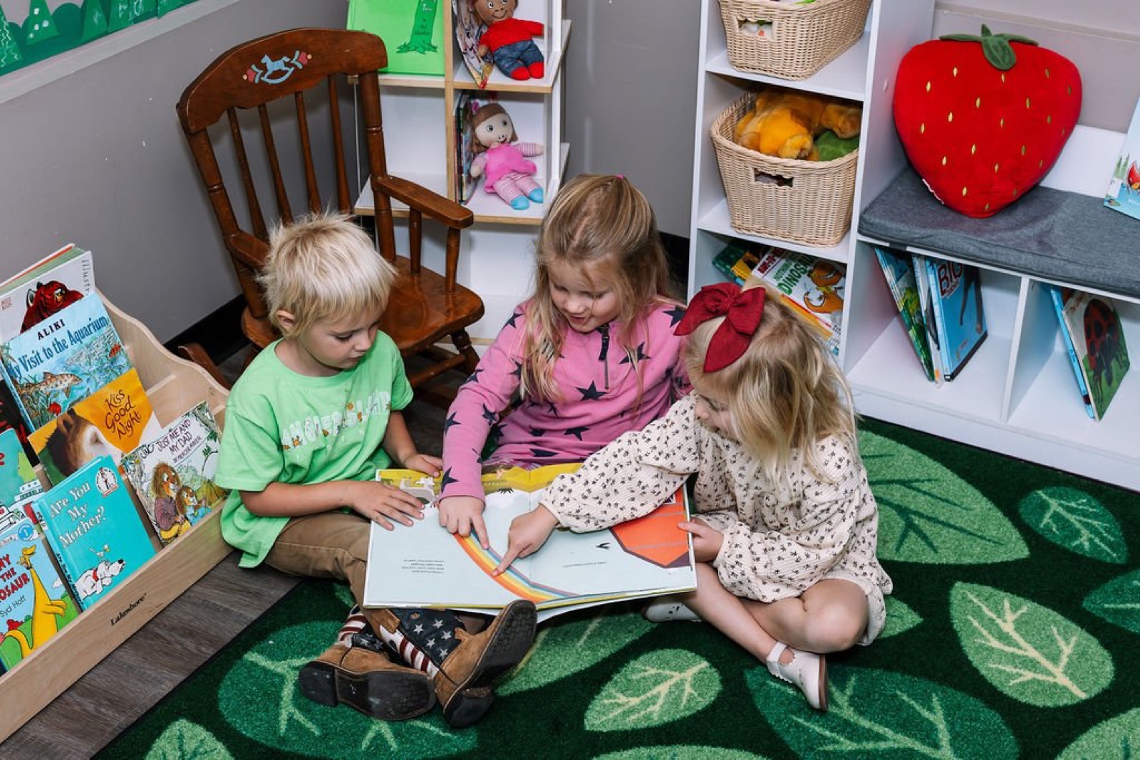 Three preschoolers reading a picture book together on a leaf-patterned rug